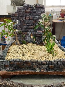 Miniaturisierte Ruine mit Pflanzen, Steinmauer und sandigem Boden.